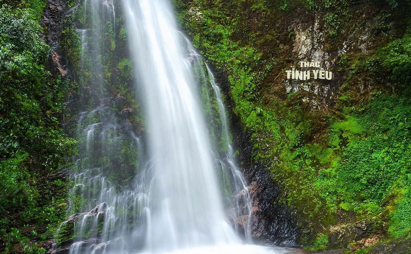 Love Waterfall dropping straight down a moss-covered rock face, next to the "Thác Tình Yêu" sign.