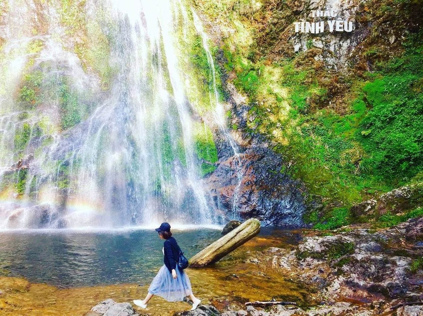 A visitor stepping across the pool at the foot of Love Waterfall, a small rainbow in the spray.