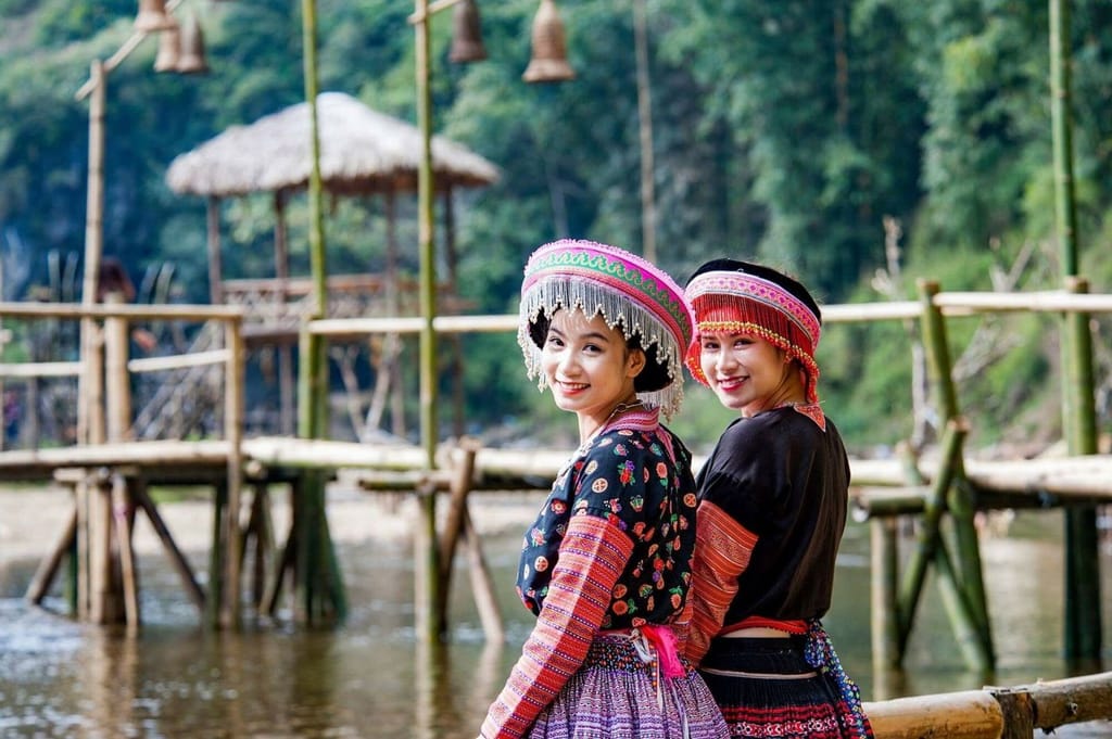 Zwei Mädchen in Hmong-Tracht posieren auf einer Bambusbrücke über dem Bach.