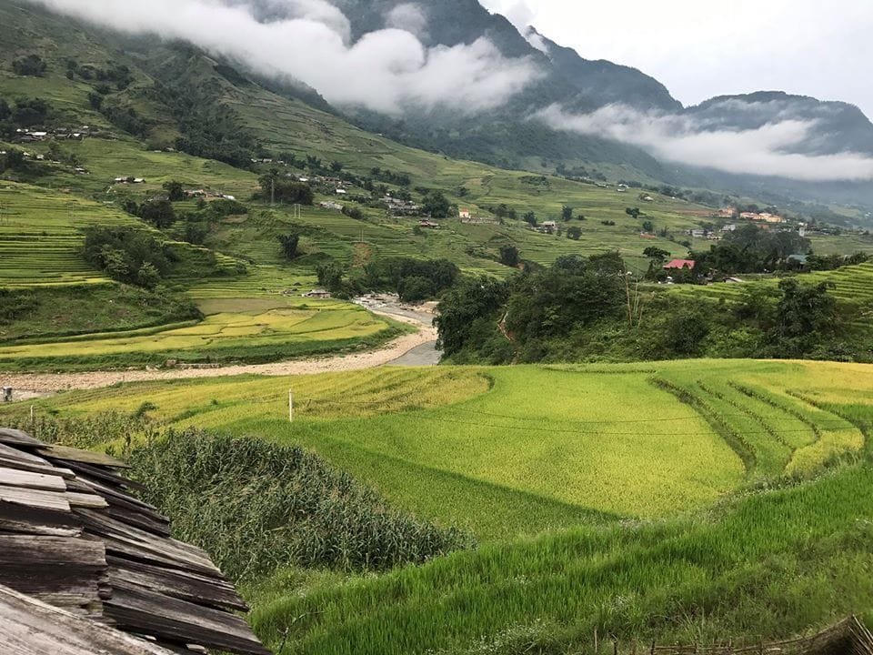 Lao Chai terraces seen past a wooden-shingle roof, with the Muong Hoa stream and clouds over the mountains beyond.