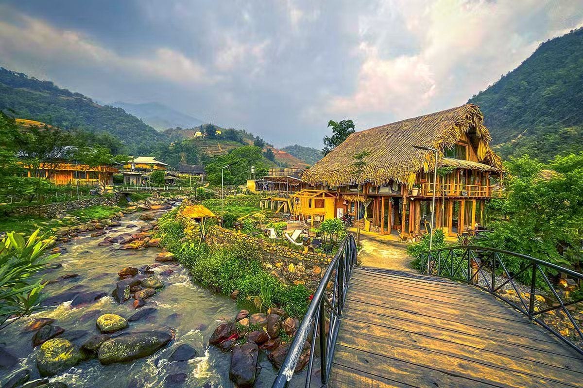 A thatched-roof homestay beside a small stream and wooden footbridge in Lao Chai.