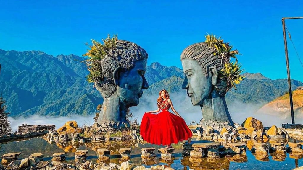 Eine Besucherin im roten Kleid zwischen den beiden einander zugewandten Kopfskulpturen am Wasser.