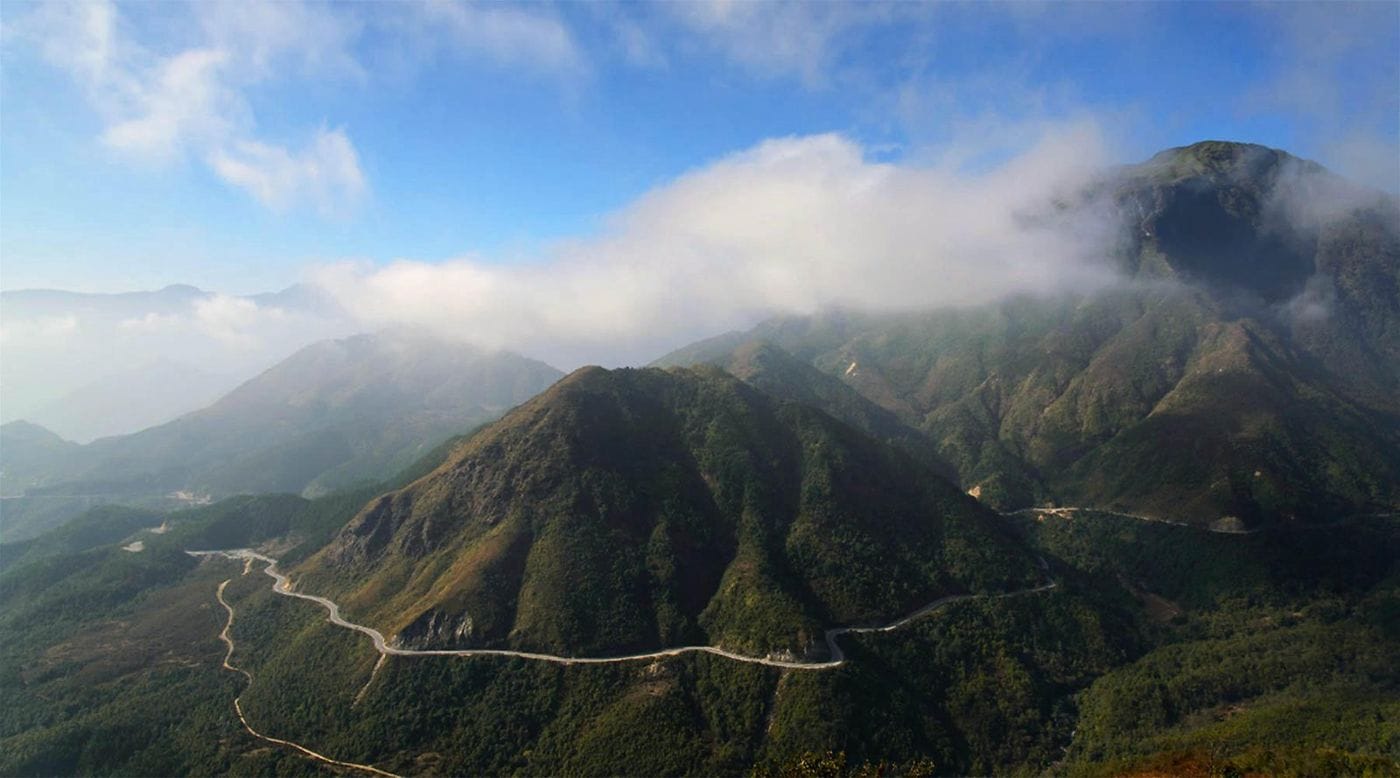 The Ô Quy Hồ road contouring along the mountainside under white clouds on the peaks.