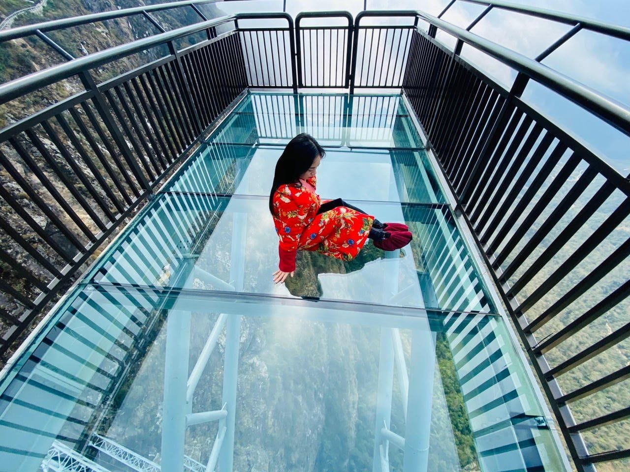 A visitor sitting on the glass deck, with the drop and forest visible below.