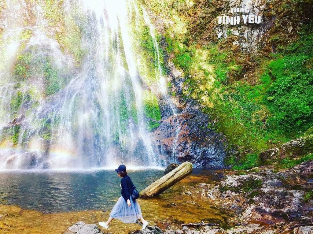 Eine Besucherin steigt über das Becken am Fuß des Liebeswasserfalls, ein kleiner Regenbogen in der Gischt.